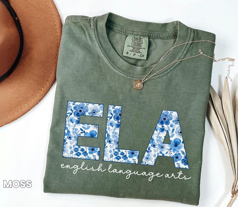 May include: Moss green t-shirt with the letters "ELA" in a blue floral pattern. The words "english language arts" are printed below the letters in a cursive font. A gold necklace is draped across the shirt.