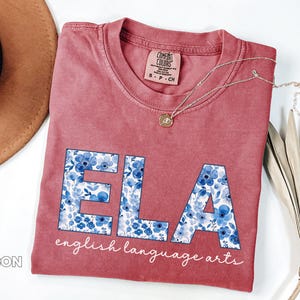 May include: Crimson-colored t-shirt with the letters "ELA" in a floral blue pattern. The words "english language arts" are written below the letters in a script font. A gold necklace and a brown hat are also in the image.