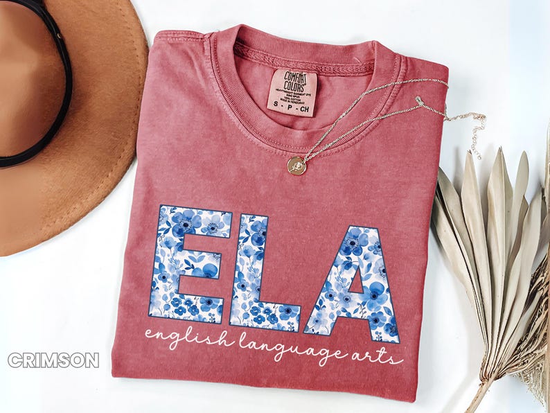 May include: Crimson-colored t-shirt with the letters "ELA" in a floral blue pattern. The words "english language arts" are written below the letters in a script font. A gold necklace and a brown hat are also in the image.
