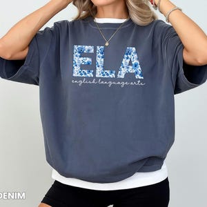 May include: Denim blue t-shirt with the letters "ELA" in a floral pattern. Below the letters, the words "english language arts" are written in a cursive font. The shirt has a relaxed fit.