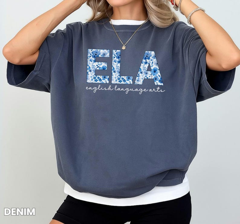 May include: Denim blue t-shirt with the letters "ELA" in a floral pattern. Below the letters, the words "english language arts" are written in a cursive font. The shirt has a relaxed fit.