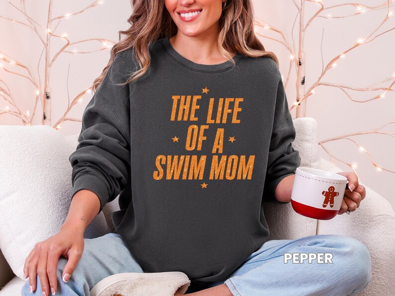 May include: Dark gray sweatshirt with the text "THE LIFE OF A SWIM MOM" in orange. The sweatshirt has a crew neck and long sleeves. A white mug with a red base and a gingerbread man design is held in the hand.