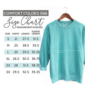 May include: A turquoise Comfort Colors sweatshirt hangs on a wooden hanger. A size chart in inches is displayed, showing measurements for width, length, and sleeve length for sizes S to 3X. The sweatshirt's length and width are indicated with white lines.