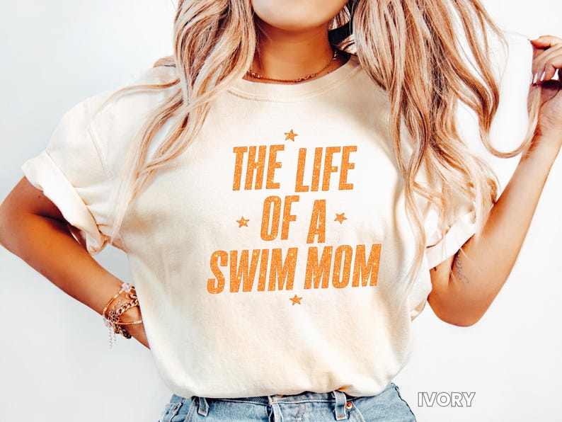 May include: Ivory-colored t-shirt with the text "THE LIFE OF A SWIM MOM" in orange, with star accents. The shirt has a crew neck and short sleeves. The word "IVORY" is printed at the bottom right.