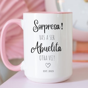 May include: White ceramic mug with a pink handle and interior. The mug features the text "Sorpresa! Vas a ser Abuelita Otra Vez!" in black script, along with a heart symbol and "EST. 2025". A charming gift for a grandmother.