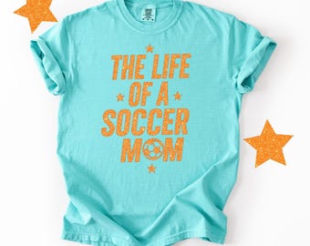 May include: A turquoise t-shirt with the text "THE LIFE OF A SOCCER MOM" in orange glitter lettering. The design includes stars and a soccer ball graphic. Two orange glitter star decorations are on the white background.