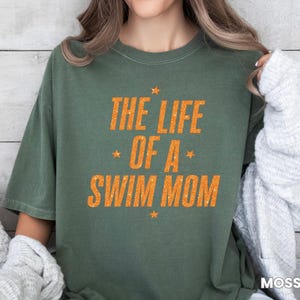 May include: Olive green t-shirt with the text "THE LIFE OF A SWIM MOM" in orange, with star accents. The shirt is a casual, comfortable style, perfect for swim team parents.