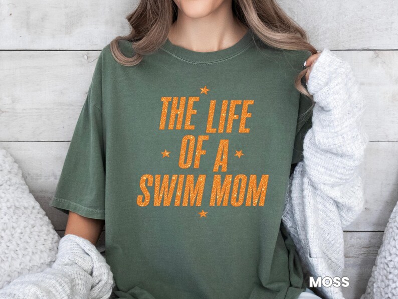 May include: Olive green t-shirt with the text "THE LIFE OF A SWIM MOM" in orange, with star accents. The shirt is a casual, comfortable style, perfect for swim team parents.