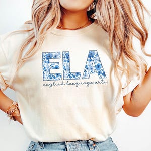 May include: Cream-colored t-shirt with the letters "ELA" in blue floral print. Below the letters, the words "english language arts" are written in a cursive font. The shirt is made of a soft material.