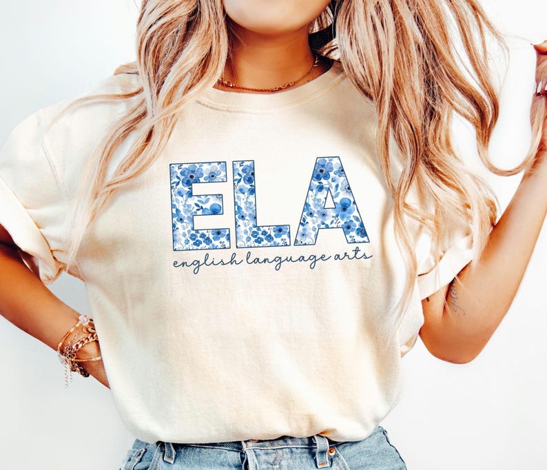 May include: Cream-colored t-shirt with the letters "ELA" in blue floral print. Below the letters, the words "english language arts" are written in a cursive font. The shirt is made of a soft material.