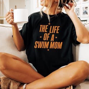 May include: Black t-shirt with the text "THE LIFE OF A SWIM MOM" in orange, with star accents. The shirt is worn by a person sitting on a couch and holding a white mug.