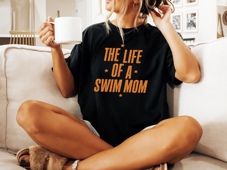 May include: Black t-shirt with the text "THE LIFE OF A SWIM MOM" in orange, with star accents. The shirt is worn by a person sitting on a couch and holding a white mug.