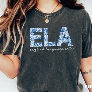 May include: Dark gray t-shirt with the letters "ELA" in a blue floral pattern. The words "english language arts" are written below the letters in a cursive font. The shirt is a casual style.