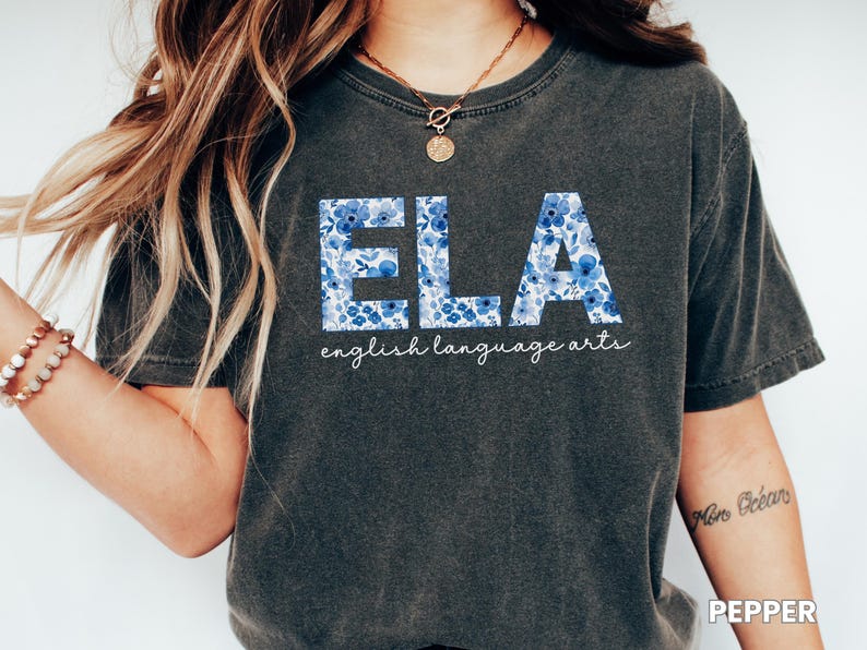 May include: Dark gray t-shirt with the letters "ELA" in a blue floral pattern. The words "english language arts" are written below the letters in a cursive font. The shirt is a casual style.