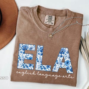 May include: Brown t-shirt with the letters "ELA" in blue floral print, with the words "english language arts" below. A silver necklace with a pendant is draped across the shirt. The shirt is a Comfort Colors brand.