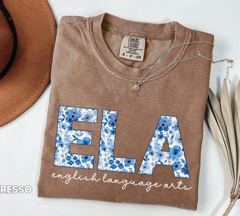 May include: Brown t-shirt with the letters "ELA" in blue floral print, with the words "english language arts" below. A silver necklace with a pendant is draped across the shirt. The shirt is a Comfort Colors brand.