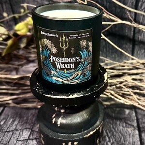 May include: A black candle jar with a blue and gold label depicting a trident and waves. The label reads "Poseidon's Wrath" and lists the scent notes: Perigrain, Sea Salt, Aloe, Bamboo, Lime, Dark Musk.