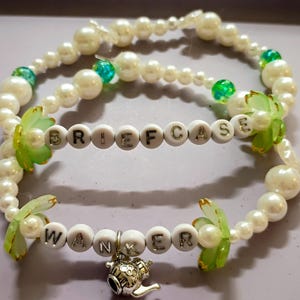 May include: Two beaded bracelets featuring white faux pearls, translucent green floral-shaped beads, and small green spherical beads. One bracelet displays the word BRIEFCASE in white letter beads, while the other features WANKER and a silver teapot charm.
