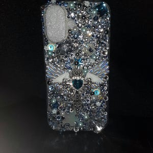 May include: A clear phone case decorated with silver, blue, and white rhinestones, pearls, and a silver cross with blue wings and a blue gem in the center.