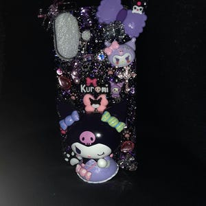 May include: A black phone case with a glittery purple background. The case features a 3D character of Kuromi, a black cat with a skull on her head, wearing a purple dress and pink bows. The case also has a purple bow with a black skull on top.