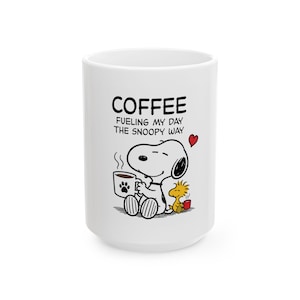 Może przedstawiać: Biały ceramiczny kubek z czarnym rysunkiem liniowym przedstawiającym Snoopy'ego trzymającego filiżankę kawy z odciskiem łapy, siedzącego obok Woodstocka. Tekst brzmi COFFEE FUELING MY DAY THE SNOOPY WAY, z małym czerwonym serduszkiem.