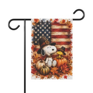 May include: A rectangular garden flag hanging on a black metal stand. The flag features a watercolor-style illustration of Snoopy and Woodstock wearing pilgrim hats, sitting among pumpkins and autumn leaves against a backdrop of the American flag.