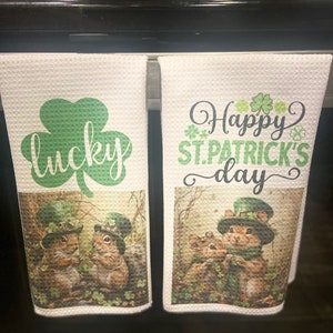 May include: Two white kitchen towels with a St. Patrick's Day theme. One towel features a green shamrock with the word "lucky" and the other towel features a green shamrock with the text "Happy St. Patrick's Day". Both towels feature a cartoon image of two squirrels wearing green hats.