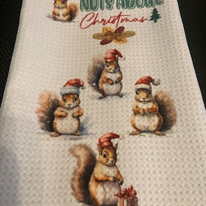 May include: A white kitchen towel with a red, green, and brown design. The towel features four squirrels wearing Santa hats and the text "NUTS ABOUT CHRISTMAS".