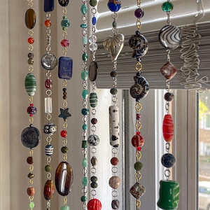 Pode incluir: Cortina decorativa de contas com uma variedade de contas coloridas de diferentes formas e tamanhos. As contas são enfiadas com correntes douradas. A cortina inclui contas em tons de azul, verde, vermelho, castanho e branco, criando um visual único e artístico.