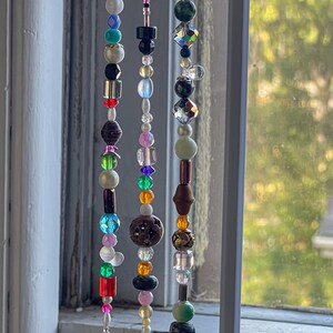 May include: A collection of decorative beaded strands, featuring a variety of colourful beads. The beads are in shades of blue, pink, green, brown, and black, with different shapes and textures. The strands hang in front of a window, catching the light.