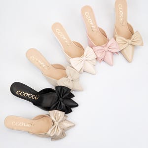 May include: A collection of pointed-toe mules in various colors, including black, pink, and beige, each adorned with a large bow. The word "ccocci" is printed in gold on the insole of each shoe. These elegant slip-ons are ideal for special occasions.