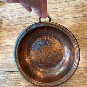 Könnte beinhalten: Ein rundes Kupfersieb mit breitem Rand und einem zentralen, perforierten Boden mit zahlreichen kleinen kreisförmigen Löchern. Die Metalloberfläche weist eine verwitterte, oxidierte Patina mit verschiedenen Braun- und Metallic-Orangetönen auf.