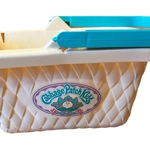 Puede incluir: Una cesta de plástico color crema con textura trenzada y un asa de color turquesa brillante. La parte frontal presenta una pegatina ovalada con el texto Cabbage Patch Kids y el gráfico de un pequeño personaje entre hojas de col verdes.