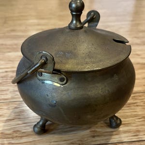 May include: A small, antique brass sugar bowl with a lid and handle. The bowl has a rounded shape, three small feet, and a hinged lid with a knob. The metal has a tarnished, aged appearance, suggesting it is vintage.