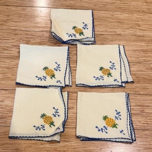 Puede incluir: Cinco servilletas de tela de color blanco roto con forma cuadrada, que presentan piñas amarillas bordadas con hojas verdes y detalles florales azules. Cada servilleta tiene un borde festoneado con costura azul, dispuestas en grupo.