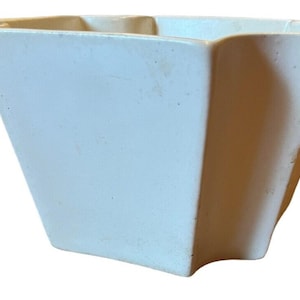 Puede incluir: Una maceta de cerámica de color crema con un borde distintivo en forma de estrella o acanalado y una base cuadrada cónica. El recipiente presenta un acabado mate suave con una textura superficial sutil y una parte superior abierta.