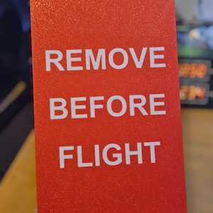 May include: A red rectangular sign with the white text "REMOVE BEFORE FLIGHT". The sign has a textured surface and is likely used in aviation or aerospace applications.