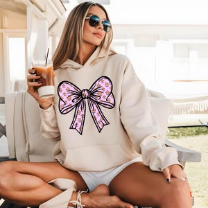 May include: A cream-colored hoodie with a large pink bow printed on the front. The bow is made up of smaller pink bows with a gold glitter pattern.