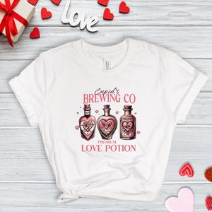 以下が含まれることがあります： 「Cupid's Brewing Co. Premium Love Potion」とラベルが貼られた3本のボトルがピンク色のグラフィックで描かれた白いTシャツです。