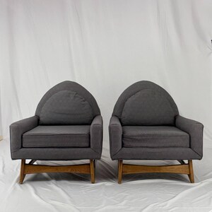 May include: Two gray armchairs with arched backs and wooden legs. The chairs have a boxy shape with a cushioned seat and back. The wooden legs are a light brown color, and the fabric appears to be a textured gray.