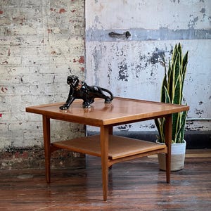 May include: A mid-century modern wooden coffee table with a lower shelf and a black panther figurine. The table has a square top and tapered legs. The panther has a gold collar. A tall potted plant is in the background.
