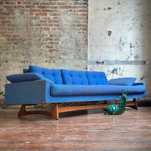 May include: A mid-century modern blue sofa with a wooden base and legs. The sofa has a tufted back and armrests. A green ceramic snail figurine sits on the wooden floor in front of the sofa.