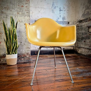 May include: A mid-century modern yellow chair with a curved back and armrests. The chair has a chrome base with four legs. A potted snake plant is in the background. The chair is on a wooden floor.