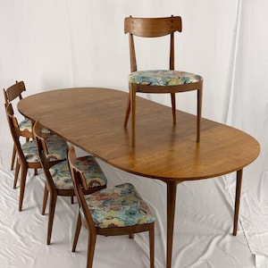 May include: A mid-century modern dining set featuring a large oval wooden table and six wooden chairs. The chairs have floral patterned seat cushions. The table and chairs are made of a light brown wood.