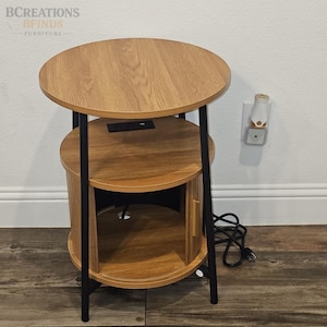 May include: A round side table with a light brown wood grain finish, featuring a top surface, a middle shelf with an integrated black power outlet, and a bottom cabinet section with a vertical door handle, supported by four thin black metal legs.