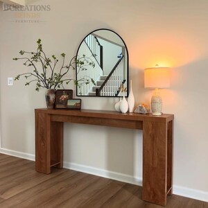 May include: A rectangular wooden console table with a smooth finish and open side shelving. The table is topped with a vase of green branches, two framed pictures, three white ceramic vases, a crystal cluster, and a table lamp with a white shade.