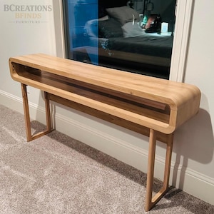 May include: A light-toned wooden console table featuring a long, rectangular top with rounded edges and a hollow open shelf running through the center. The piece is supported by four thin, straight wooden legs that extend to the floor.
