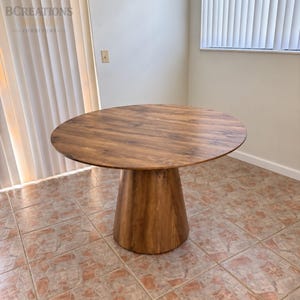 May include: A round dining table with a medium brown wood grain finish. The table features a smooth, circular top supported by a single, wide, tapered pedestal base that flares outward toward the floor, creating a stable, unified structure.