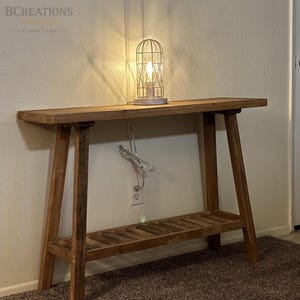 May include: A rectangular wooden console table with a natural brown finish, featuring four angled legs and a slatted lower shelf. A white wire-frame cage lamp with a glowing light bulb sits on the tabletop, with a white power cord plugged into the wall.