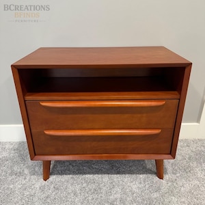 May include: A medium-brown wooden nightstand featuring an open top shelf and two drawers below. Each drawer is equipped with a long, horizontal, integrated wooden handle. The piece is supported by four tapered legs and has a smooth, finished surface.